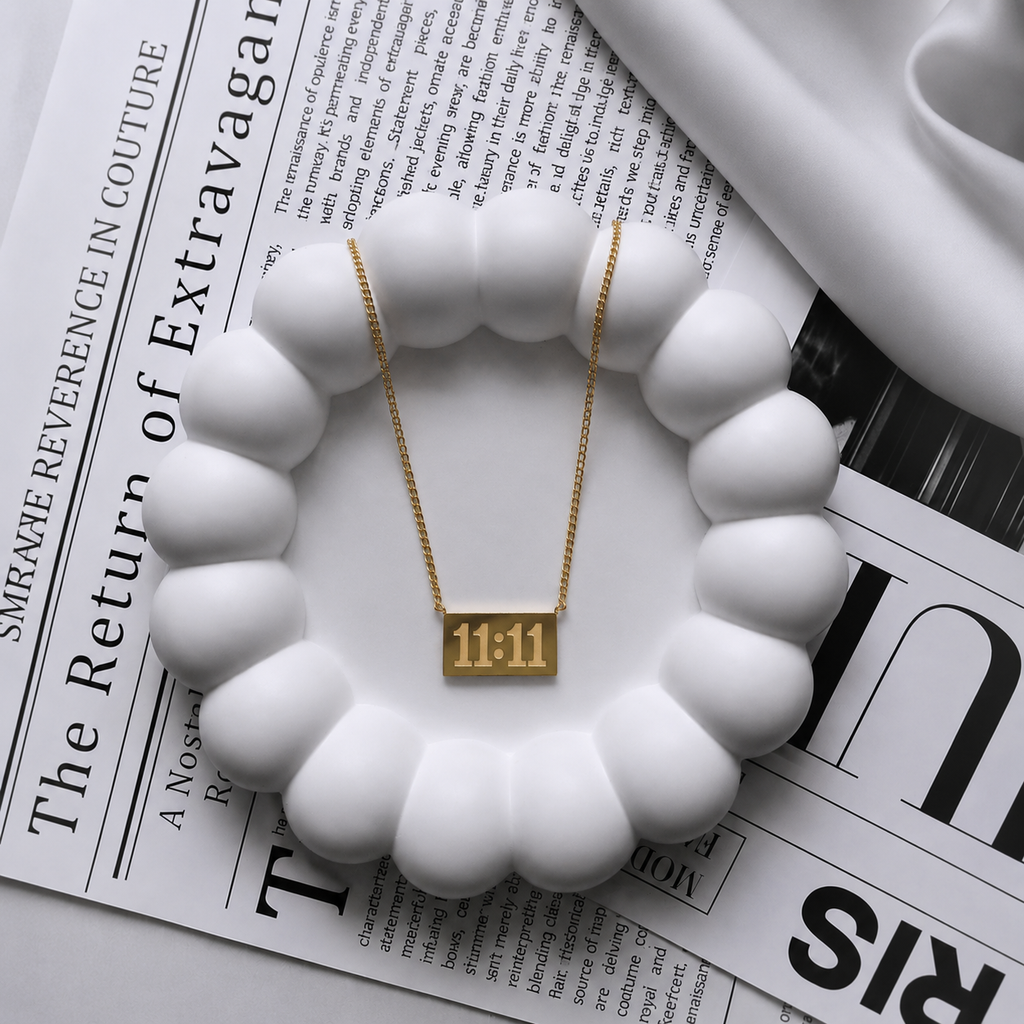 A gold rectangular pendant necklace with the numbers '11:11' on a delicate gold chain, displayed on a white scalloped dish over a black-and-white newspaper.