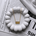 A gold rectangular pendant necklace with the numbers '11:11' on a delicate gold chain, displayed on a white scalloped dish over a black-and-white newspaper.