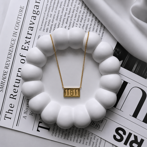 A gold rectangular pendant necklace with the numbers '11:11' on a delicate gold chain, displayed on a white scalloped dish over a black-and-white newspaper.