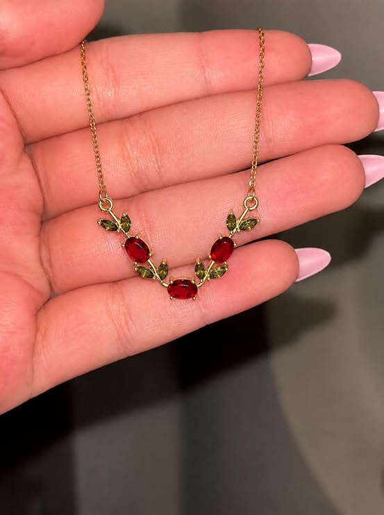 A delicate gold chain necklace with five oval red gemstones and small green leaf accents, held in a person's hand with pink manicured nails.