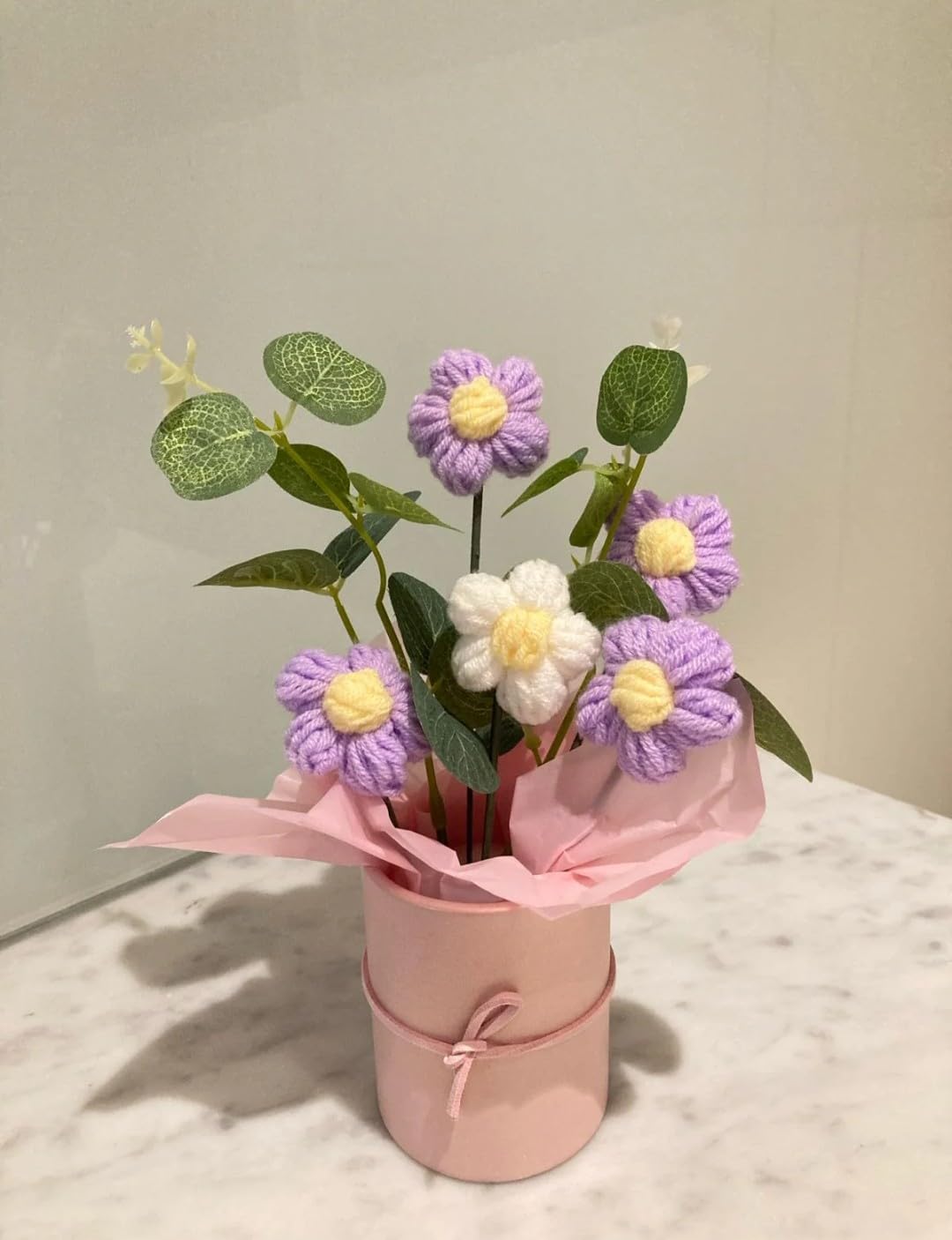 Bouquet of purple knitted daisy flowers in a pink vase on a marble surface.