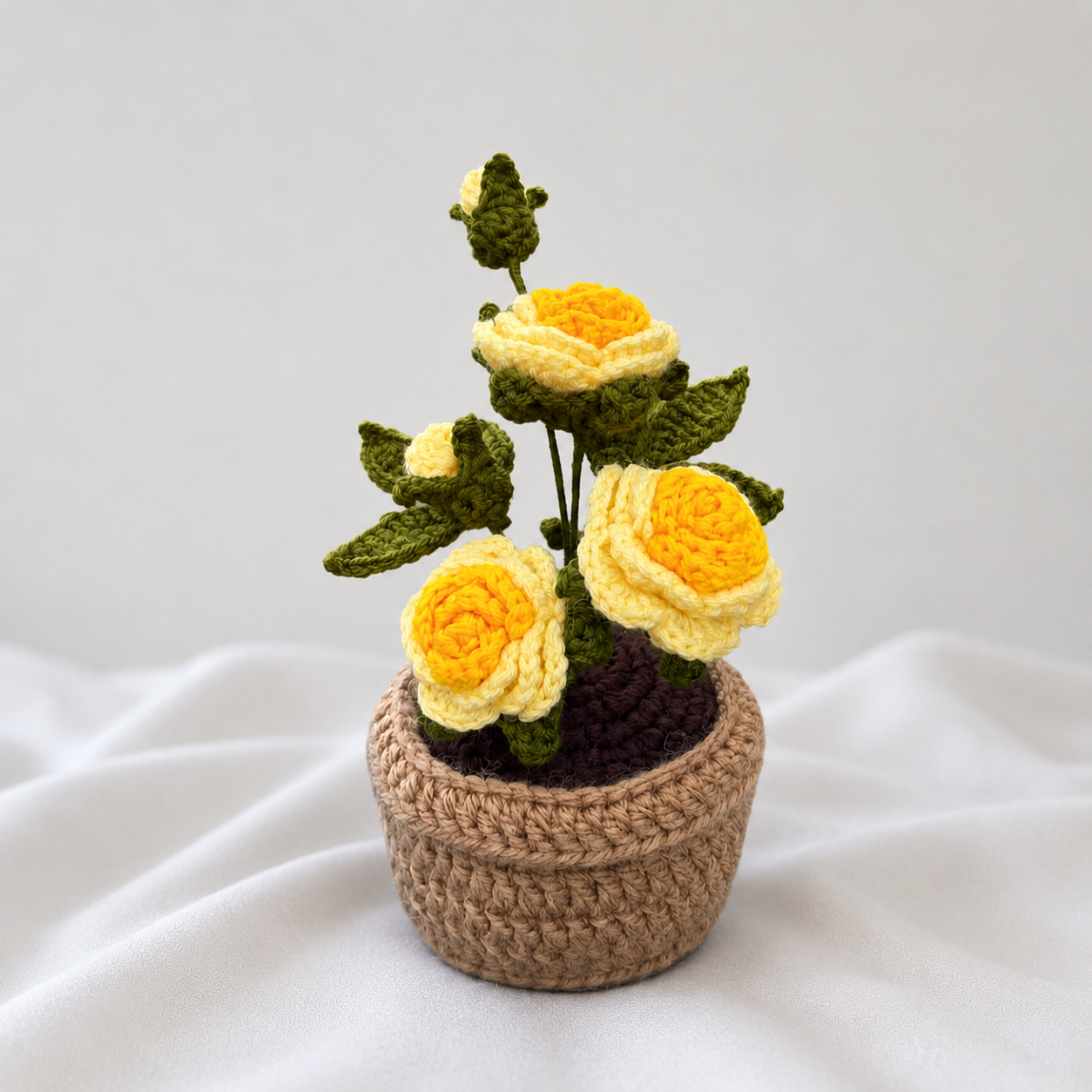 A crocheted artificial flower arrangement featuring three yellow and cream roses with green leaves, presented in a small brown woven pot on a soft white fabric background.