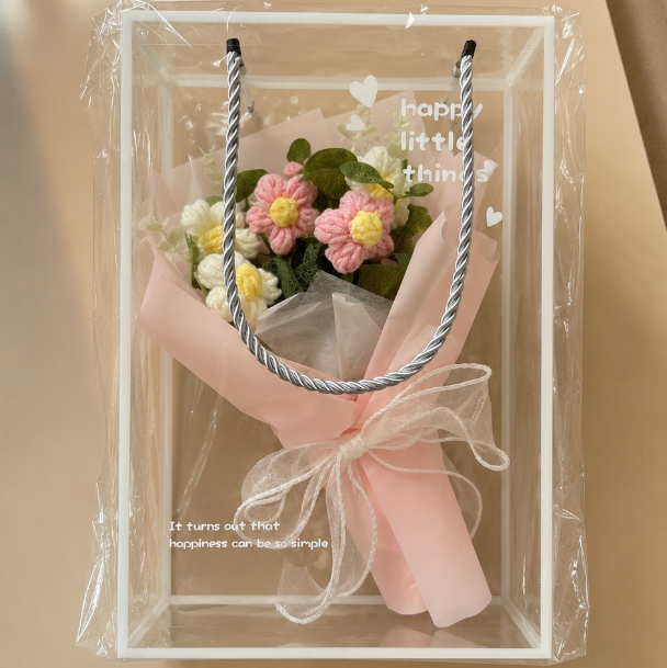 A clear acrylic gift box with a pink ribbon and small floral arrangement, photographed on a warm beige background with soft window shadows.