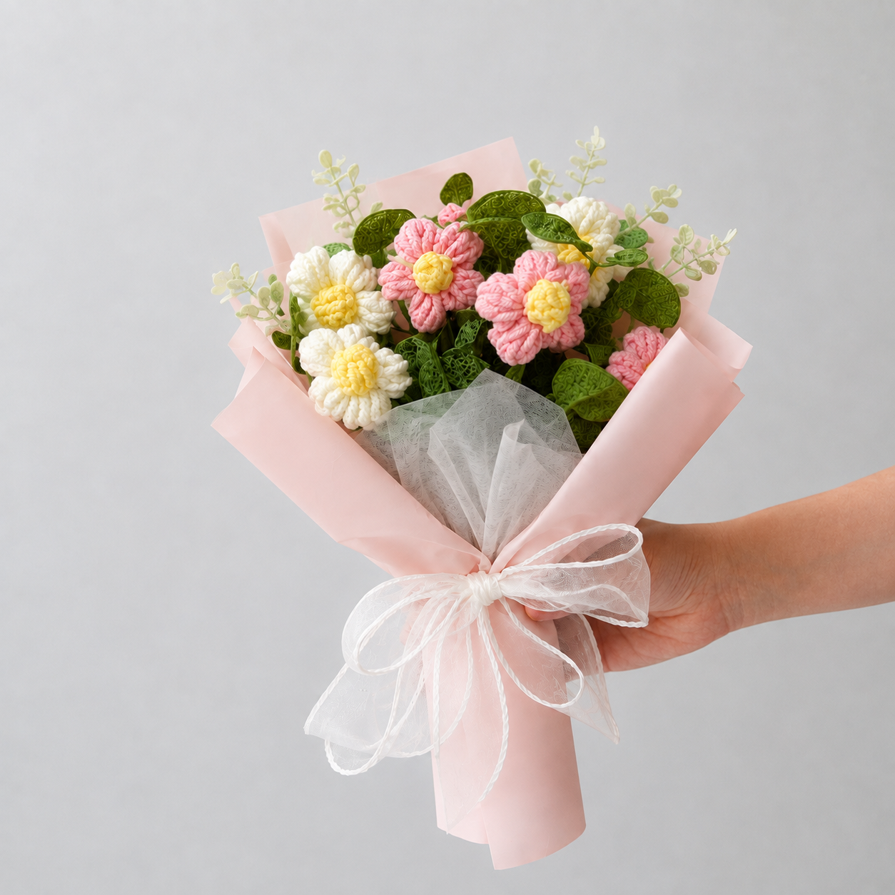 A hand holding a small bouquet of crocheted flowers wrapped in light pink paper with a white tulle ribbon, against a plain light gray background.