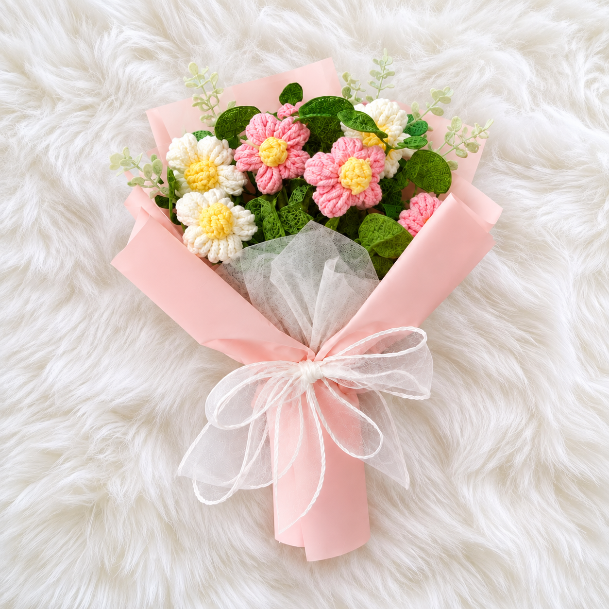 A handmade crochet flower bouquet wrapped in light pink paper and tied with a white sheer ribbon, placed on a white faux fur background.