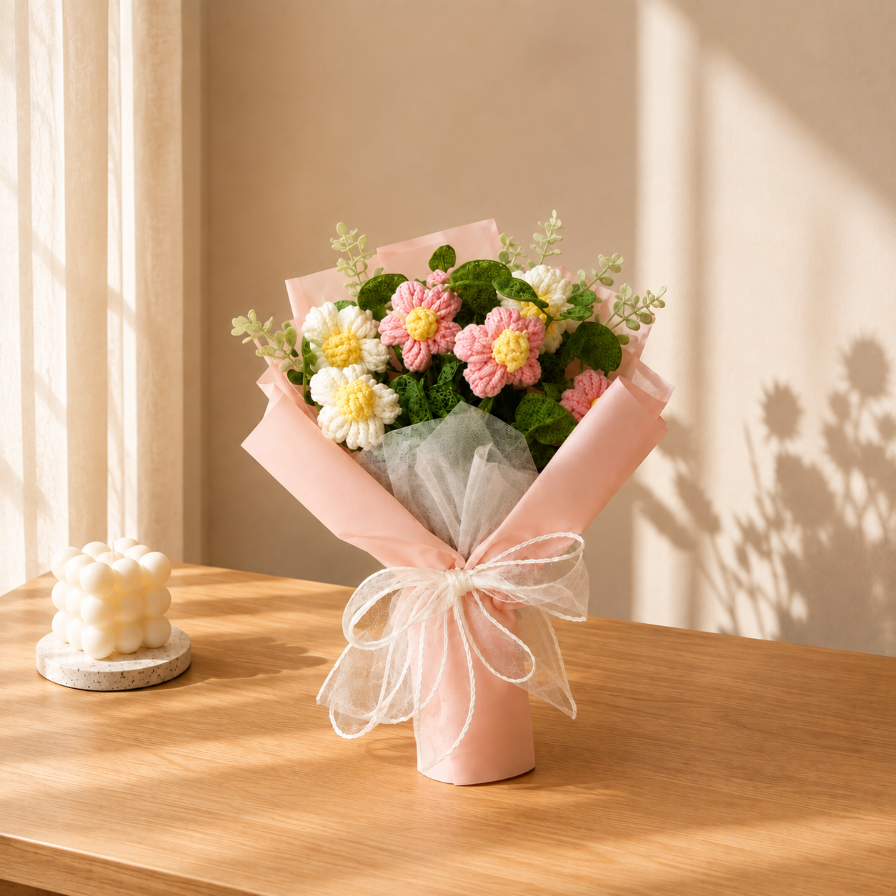 A small bouquet of pink, white, and yellow flowers wrapped in blush pink paper and tied with a sheer white ribbon, displayed on a wooden table in a softly lit room.
