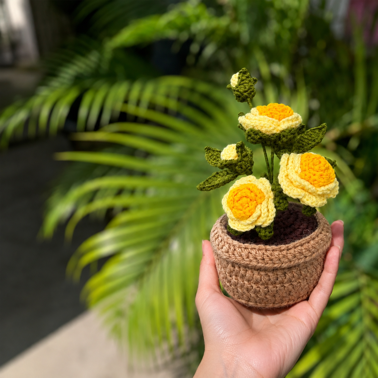 A hand holds a small crocheted flower pot with three yellow and cream crochet roses and green leaves, set against a blurred background of green palm leaves.