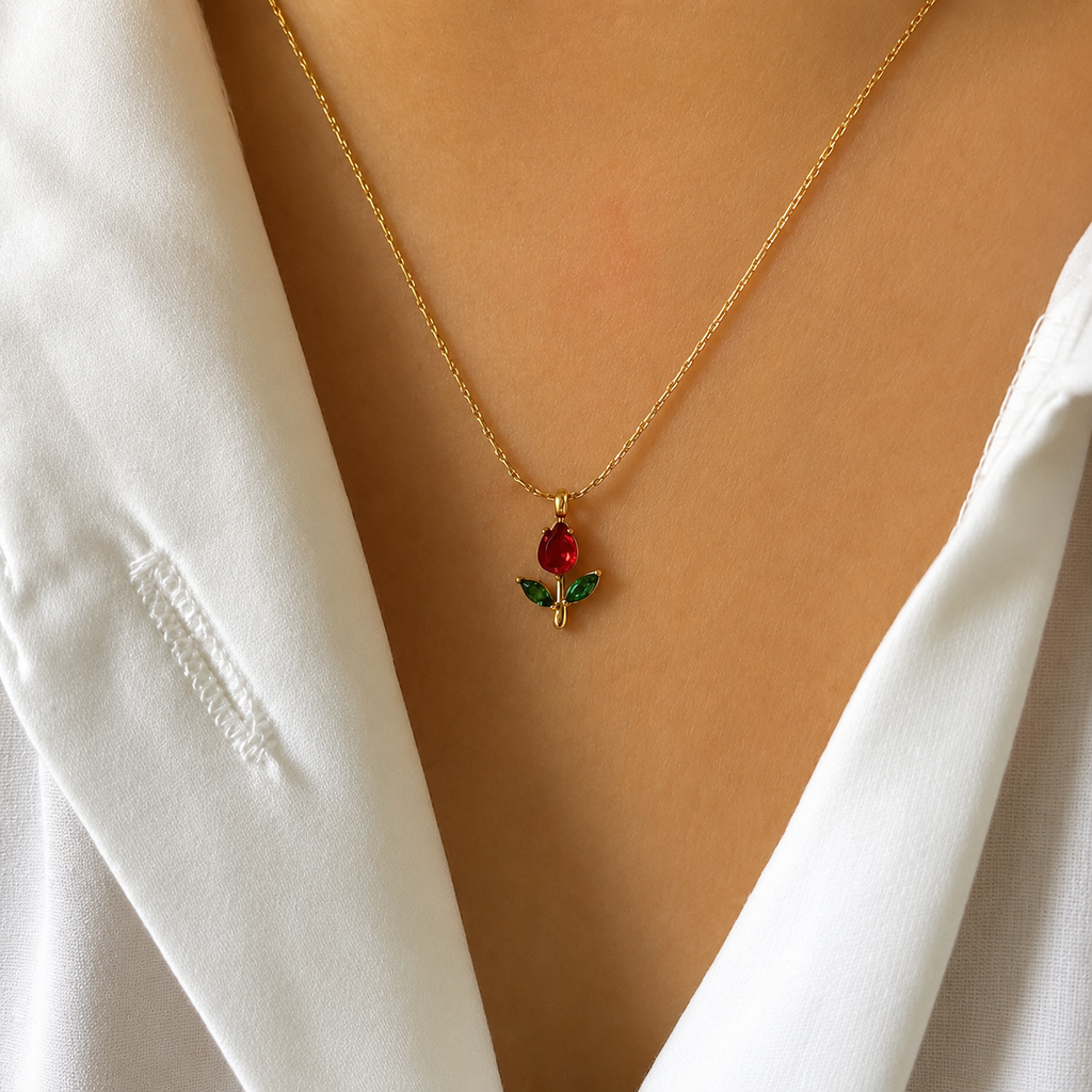 A close-up of a person wearing a delicate gold chain necklace with a small tulip pendant featuring a red gemstone and green enamel leaves, styled with a white collared shirt.