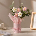 A pink cylindrical flower box wrapped with a pink ribbon, filled with pink and white artificial flowers and green stems, styled on a tabletop with a cup and framed print in the background.