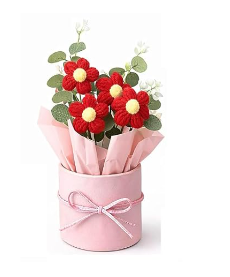 A pink cylindrical gift box wrapped with a pink ribbon, filled with red crocheted flowers with yellow centers and green leaves, set against a plain white background.