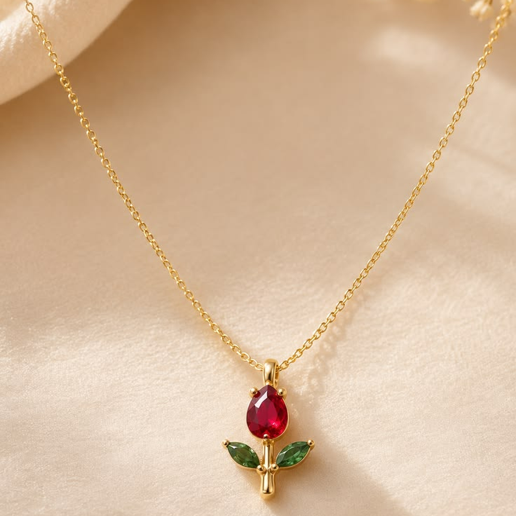 A delicate gold chain necklace featuring a pendant shaped like a tulip flower, with a teardrop-shaped red gemstone center and two green leaf-shaped stones, displayed on a soft beige fabric background.